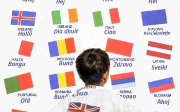 EIFA A person stands with their back to the camera facing an International Open Day wall covered in international flags, each labeled with the country name and a greeting in the local language.