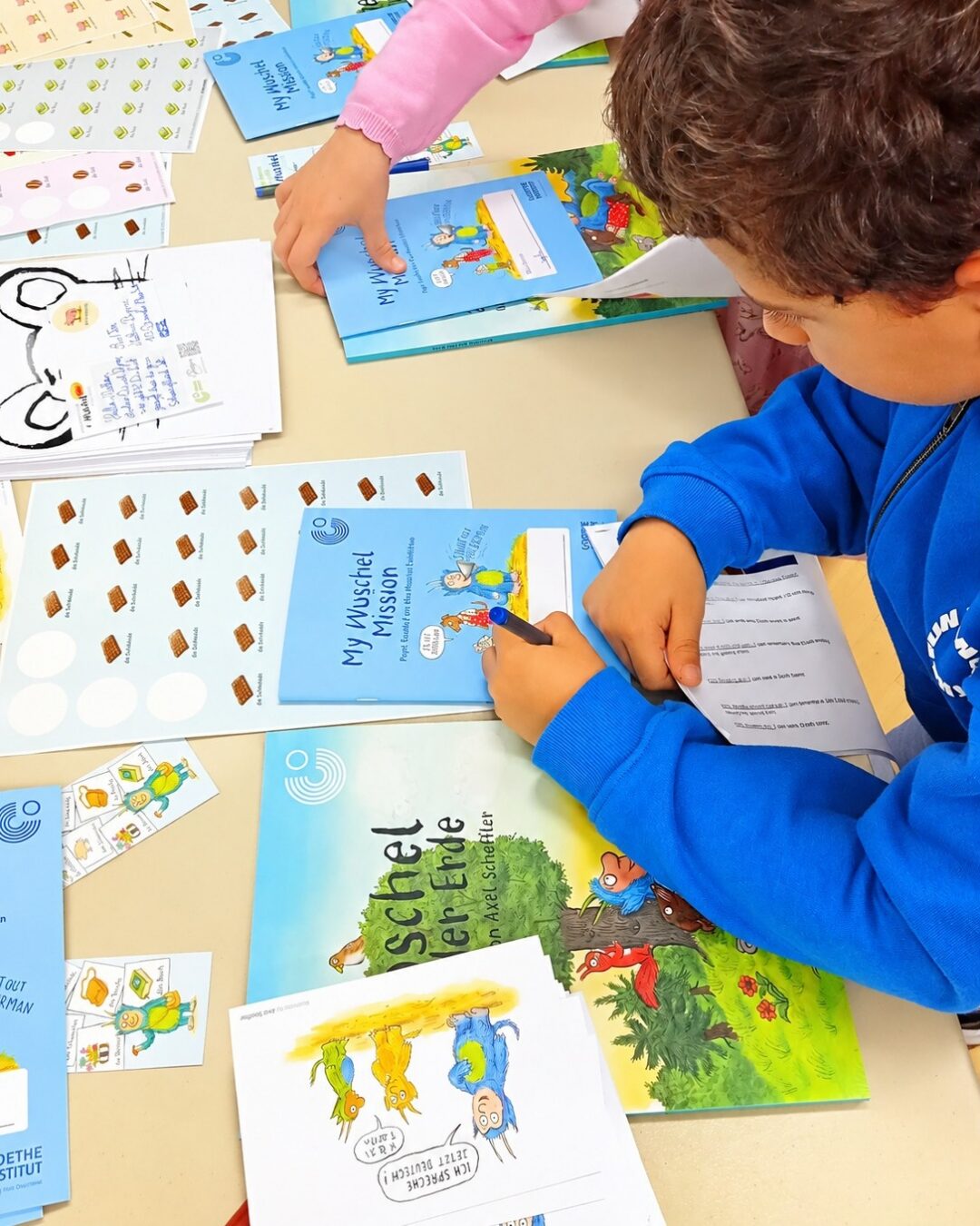 EIFA Un enfant vêtu d'un pull bleu écrit dans un cahier d'exercices My Wuschel Mission à une table jonchée de matériel pédagogique, d'autocollants et de livres d'histoires illustrés.