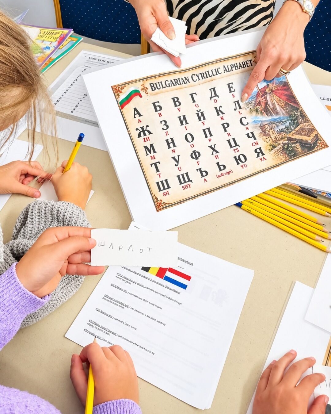 EIFA Un enseignant montre du doigt une affiche représentant l'alphabet cyrillique bulgare tandis que les élèves s'entraînent à écrire sur des fiches d'exercices, assis à une table.
