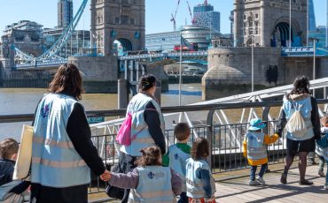 EIFA Un groupe d'adultes et d'enfants, vêtus de gilets assortis, profitent d'une promenade Journées découverte le long des quais près du Tower Bridge à Londres. Les tours emblématiques du pont et le paysage urbain sont magnifiquement visibles sous un ciel bleu azur.