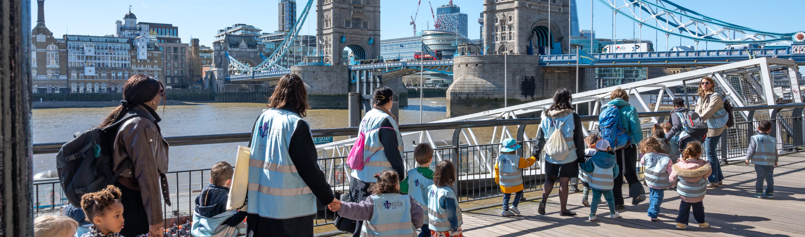 EIFA A group of adults and children wearing reflective vests enjoy a lively Taster Day as they stroll along the riverside boardwalk. In the background, the iconic Tower Bridge spans the river under a clear blue sky, creating a vibrant and memorable scene.