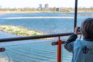 EIFA A nursery pupil in a blue vest stands on a balcony, transported into a realm of discovery as they peer through binoculars over the wide lake, surrounded by reeds and distant buildings on a clear day.