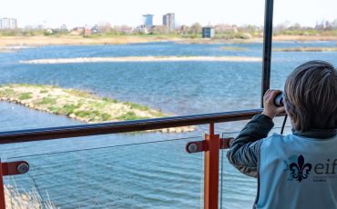 EIFA Un enfant vêtu d'un gilet bleu sarcelle avec l'inscription « eifa » dessus regarde à travers des jumelles un paysage à couper le souffle, rappelant Les PS au coeur de Londres, avec un lac, de petites îles et des bâtiments lointains vus depuis un balcon encadré de panneaux de verre.