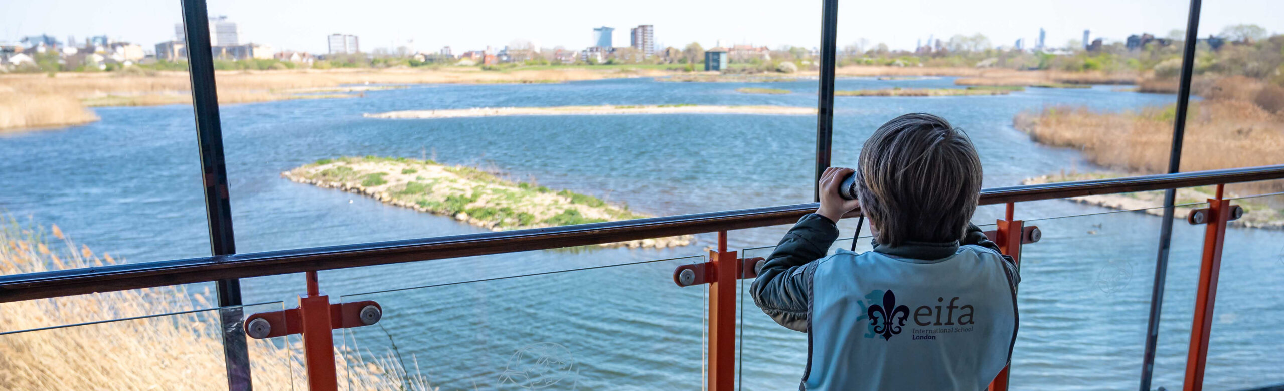 EIFA A nursery pupil in a blue vest stands on a balcony, transported into a realm of discovery as they peer through binoculars over the wide lake, surrounded by reeds and distant buildings on a clear day.