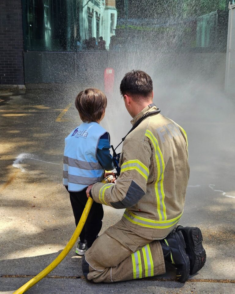 EIFA A firefighter kneels beside a young child wearing a vest, helping the child spray water from a fire hose, with water mist in the air and sunlight shining on the wet ground.