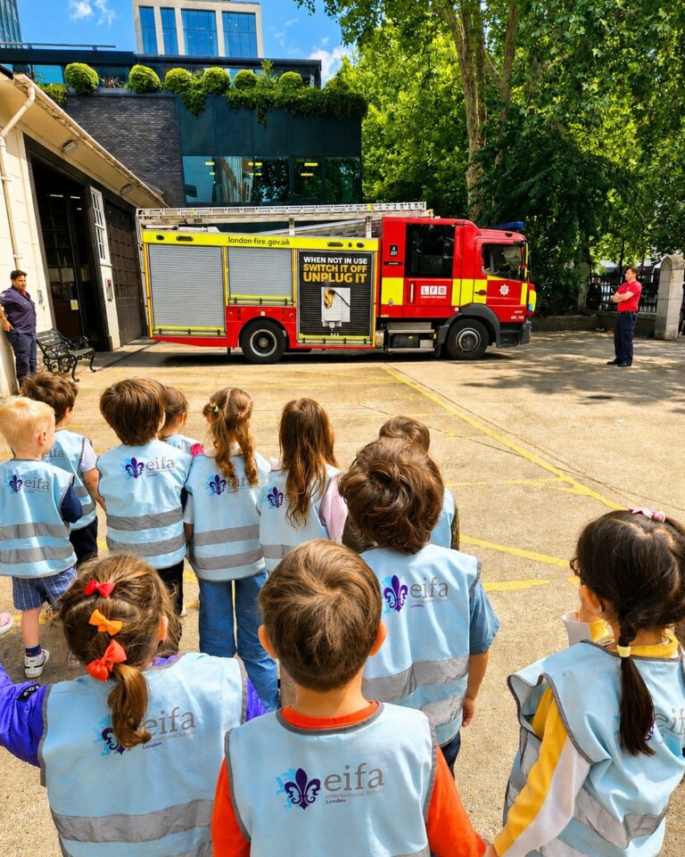 EIFA A group of young children wearing blue EIFA vests stand outside, facing a red fire truck parked near a building on a sunny day. An adult in a red shirt stands near the truck.