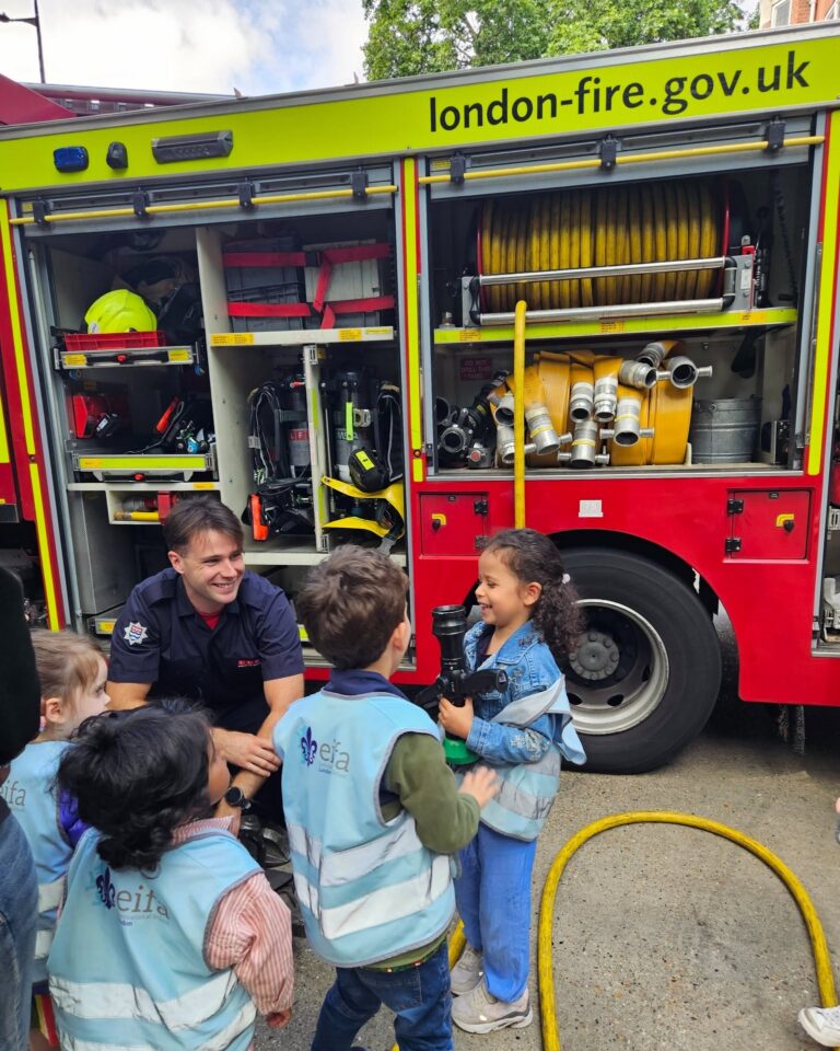 EIFA A firefighter kneels by an open fire engine, smiling and talking with young children wearing blue vests. One child holds a fire hose. The fire engine’s compartments and equipment are visible.