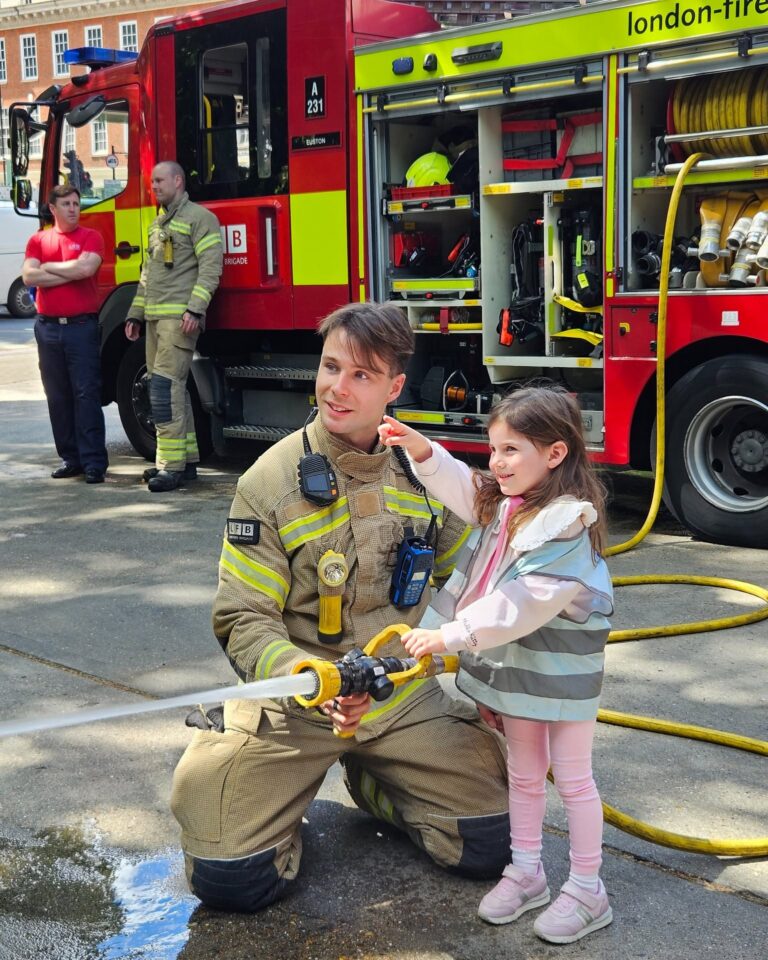 EIFA A firefighter kneels beside a smiling young girl as they hold a fire hose together. A fire engine with open compartments is behind them, with other firefighters and onlookers nearby.