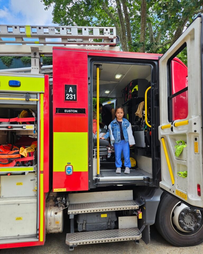 EIFA A young child in blue pants and a denim jacket stands smiling inside the open door of a red fire truck marked Euston. The trucks equipment compartments are visible, and trees are in the background.