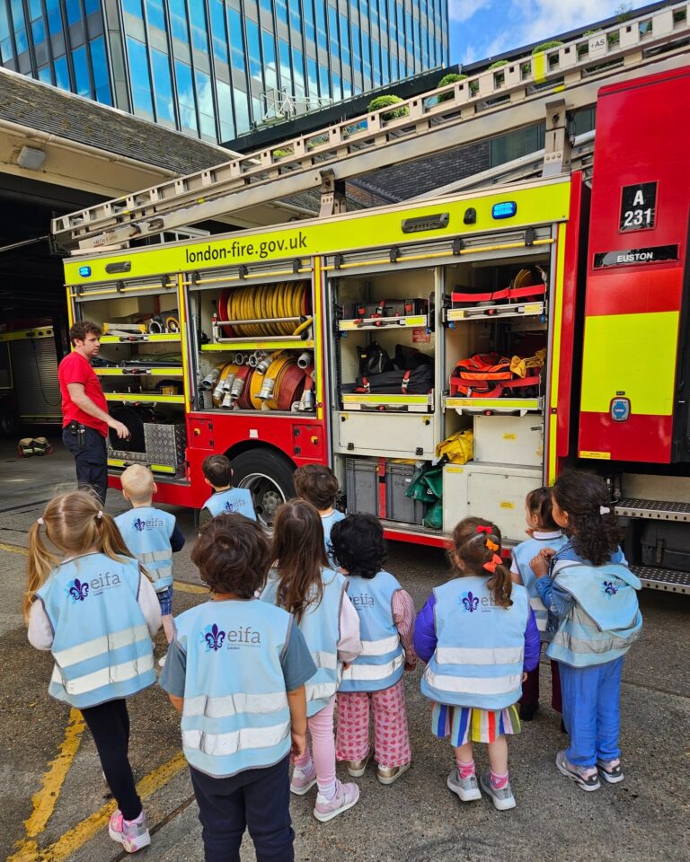 EIFA A group of young children in matching light blue vests stand in front of a fire truck, listening to a firefighter explain the equipment stored inside the vehicle. The fire truck has london-fire.gov.uk on the side.