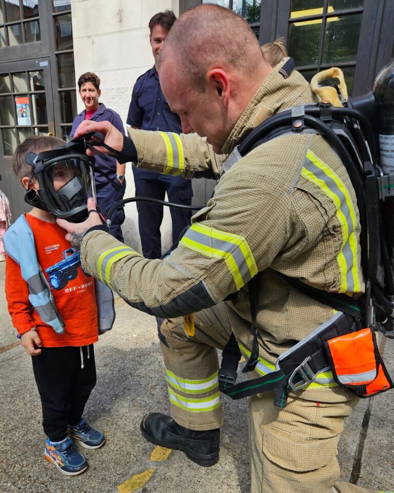 EIFA A firefighter in full gear kneels to help a young boy try on a breathing mask outside a building, while several adults and another firefighter watch in the background.