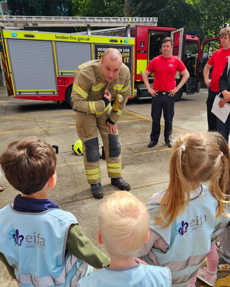 EIFA A firefighter in uniform bends down to speak with a group of young children wearing EIFA vests, while two other fire crew members and a fire engine are visible in the background.