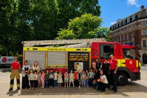 EIFA A group of children and adults stand in front of a London Fire Brigade fire truck with two firefighters, outside on a sunny day. The fire truck displays a safety message about unplugging devices.