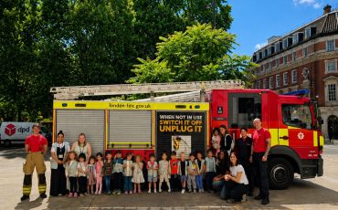 EIFA A group of children and adults stand in front of a London Fire Brigade fire truck with two firefighters, outside on a sunny day. The fire truck displays a safety message about unplugging devices.