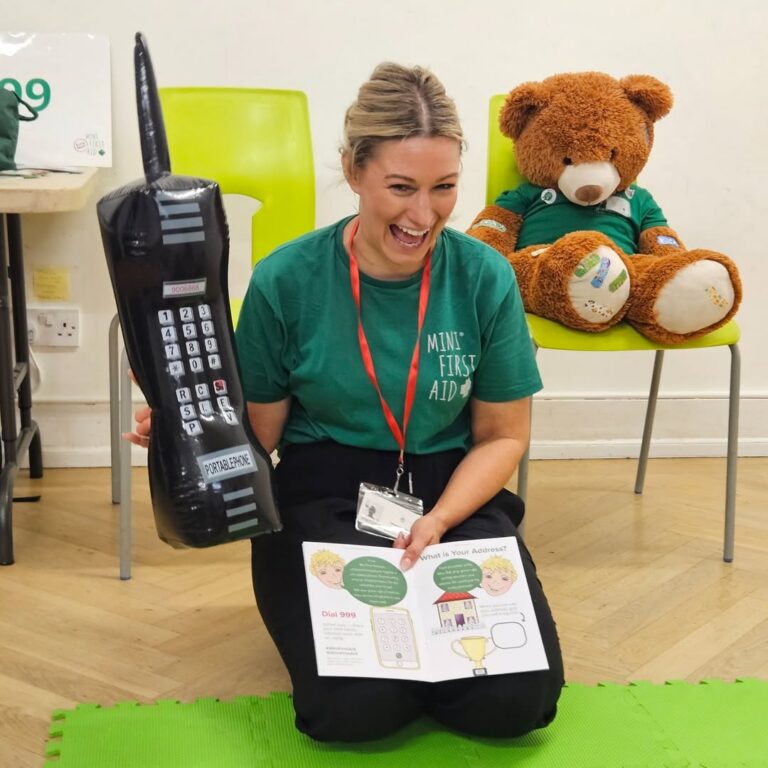 EIFA A woman kneels on the floor, smiling and holding a large inflatable phone. She wears a green Mini First Aid shirt. A teddy bear sits on a chair behind her, and she displays an open first aid booklet.