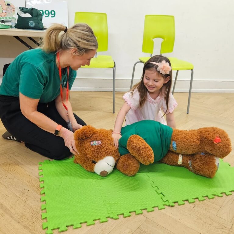 EIFA A woman and a young girl smile while practicing first aid on a large teddy bear lying on a green mat. The teddy bear has bandages and is dressed in a green shirt. Two empty chairs are in the background.