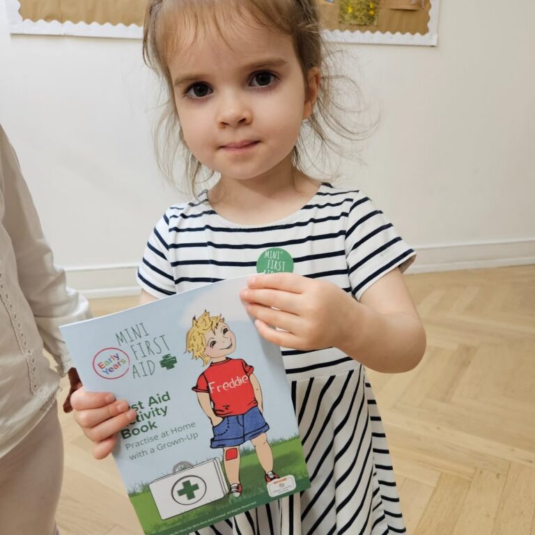 EIFA A young girl in a striped dress holds a Mini First Aid activity book and wears a matching sticker, standing indoors on a light wood floor.
