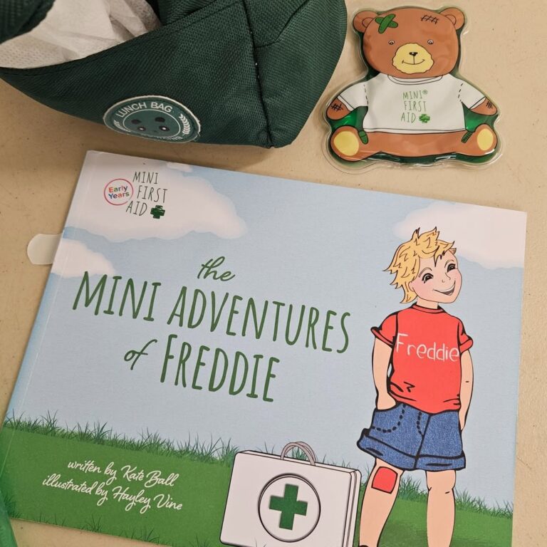 EIFA A children’s book titled The Mini Adventures of Freddie sits on a table, accompanied by a green bag and a bear-shaped cookie decorated with a white shirt that reads MINI FIRST AID.