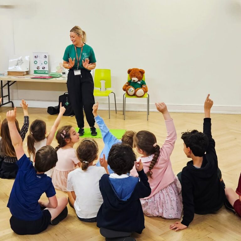 EIFA A woman speaks to a group of children sitting on the floor, many with their hands raised. Behind her are chairs, a teddy bear, and a display board, all in a bright room with a wooden floor.