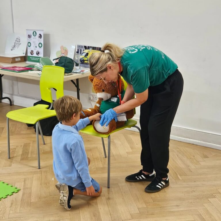 EIFA A woman in a green shirt and gloves helps a young boy bandage a large teddy bear’s leg in a classroom setting with chairs, tables, and educational materials in the background.