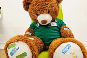 EIFA A large brown teddy bear wearing a green shirt sits on a bright yellow chair. Its paws and body are covered with colorful cartoon bandages, and it appears to be in a playful or comforting setting.