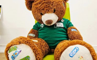 EIFA A large brown teddy bear wearing a green shirt sits on a bright yellow chair. Its paws and body are covered with colorful cartoon bandages, and it appears to be in a playful or comforting setting.