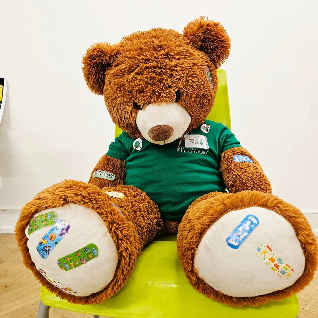 EIFA A large brown teddy bear wearing a green shirt sits on a bright yellow chair. Its paws and body are covered with colorful cartoon bandages, and it appears to be in a playful or comforting setting.