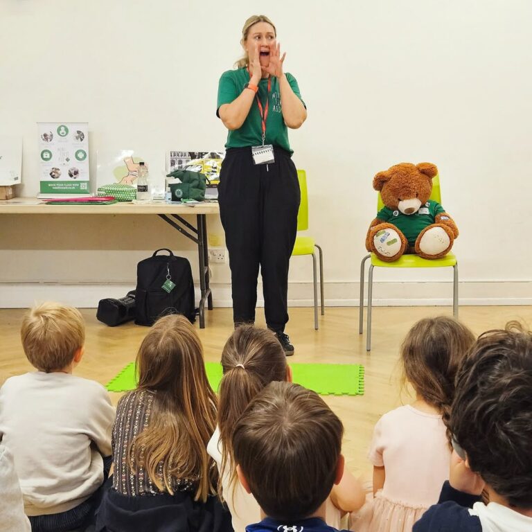 EIFA A woman stands in front of a group of seated children, holding her hands to her mouth as if calling out. A large teddy bear sits on a chair beside her, and a table with materials is in the background.