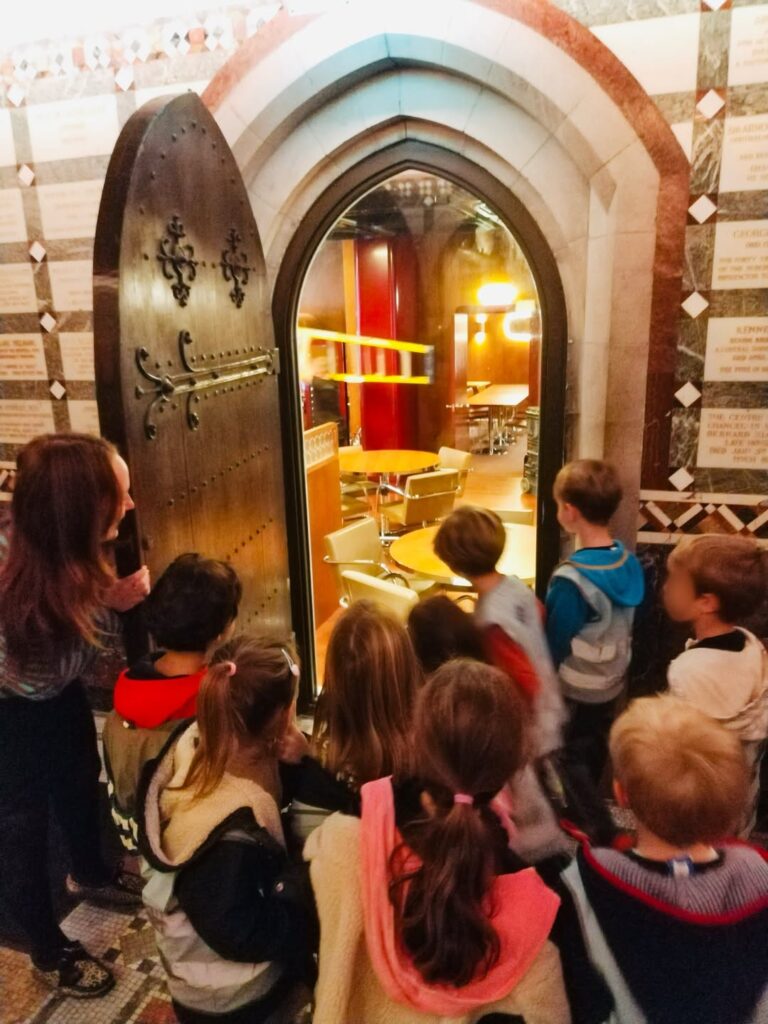 EIFA Un groupe d'enfants et un adulte se tiennent devant une porte cintrée ouverte, donnant sur une salle bien éclairée avec des tables et des chaises. Le décor semble être un bâtiment orné et carrelé.