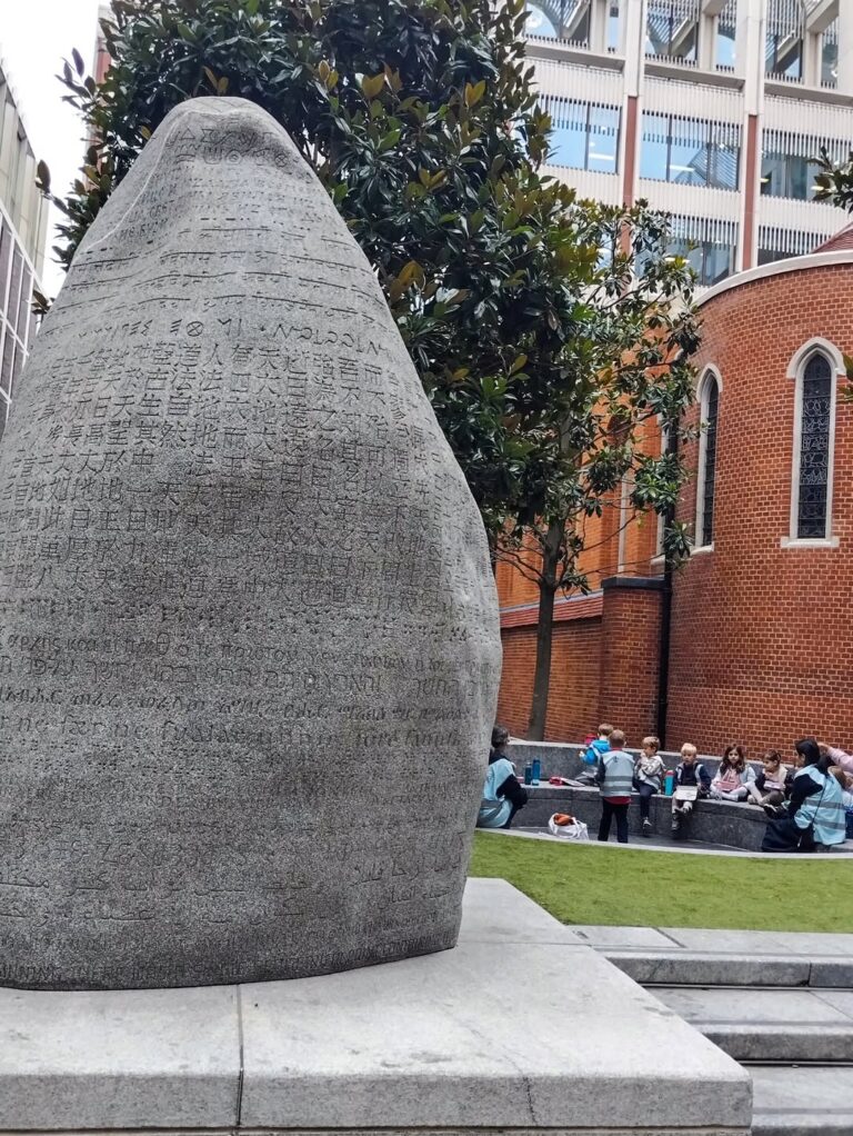 EIFA A large stone sculpture inscribed with various scripts stands in a courtyard near a brick church. In the background, people sit and talk at tables on a grassy area surrounded by buildings and trees.