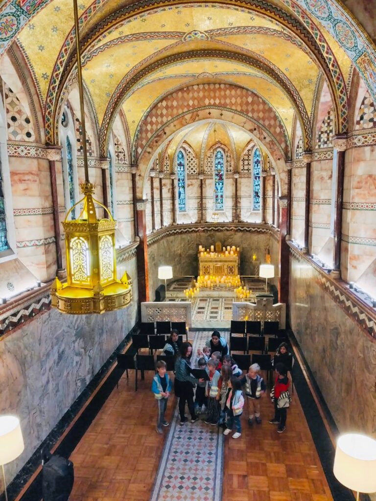 EIFA Un groupe d'enfants et d'adultes se tient dans une chapelle richement décorée, avec des plafonds voûtés et ornés, des vitraux et un autel doré. Une grande lampe suspendue se trouve au premier plan.