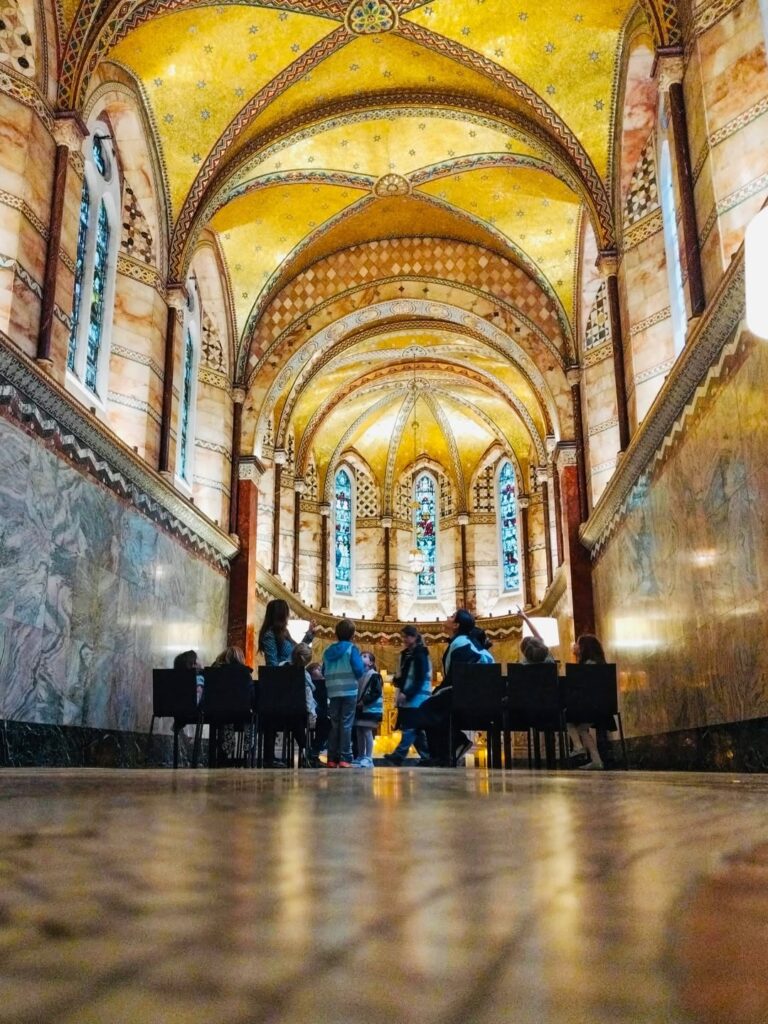 EIFA Un groupe de personnes assises et debout dans une grande salle aux plafonds voûtés dorés, aux murs de marbre et aux vitraux, vue d'un angle bas près du sol.