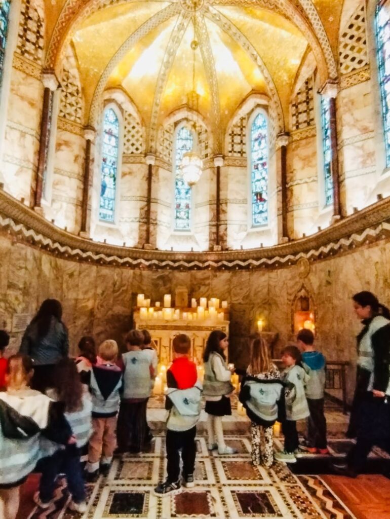 EIFA Un groupe d'enfants et deux adultes se tiennent à l'intérieur d'une chapelle ornée d'un dôme doré, de vitraux, de murs en marbre et de bougies allumées sur l'autel.