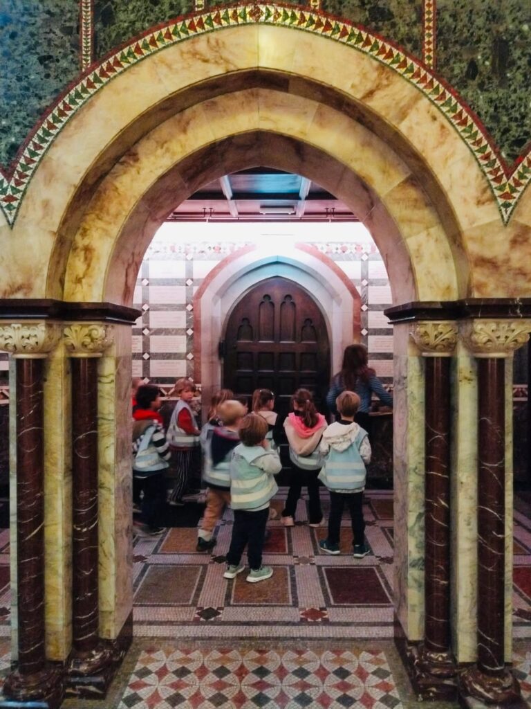 EIFA Un groupe de jeunes enfants se tient à l'intérieur d'un bâtiment aux arches ornées et aux sols à motifs, face à une grande porte en bois, accompagnés d'un adulte.