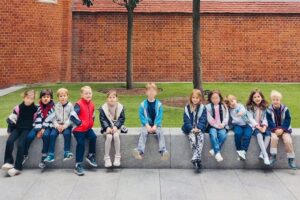 EIFA A group of eleven young children sit in a row on a low stone wall in front of a red brick building with arched windows, green grass, and two trees.
