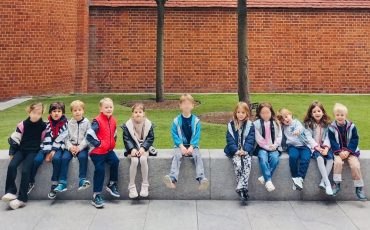 EIFA Un groupe de dix jeunes enfants est assis en rang sur un banc de pierre bas, à l'extérieur, avec des arbres, de l'herbe et un bâtiment en briques rouges à l'arrière-plan. Les enfants sont vêtus de vestes et de pantalons décontractés.
