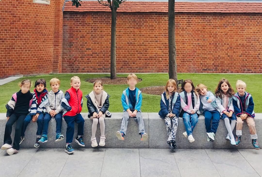 EIFA Un groupe de dix jeunes enfants est assis en rang sur un banc de pierre bas, à l'extérieur, avec des arbres, de l'herbe et un bâtiment en briques rouges à l'arrière-plan. Les enfants sont vêtus de vestes et de pantalons décontractés.