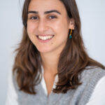 EIFA A smiling woman with long brown hair, wearing a gray sweater vest over a white shirt, stands against a light background. Her earrings reflect her friendly expression, exuding the warmth of someone who thrives in a dedicated and experienced team.