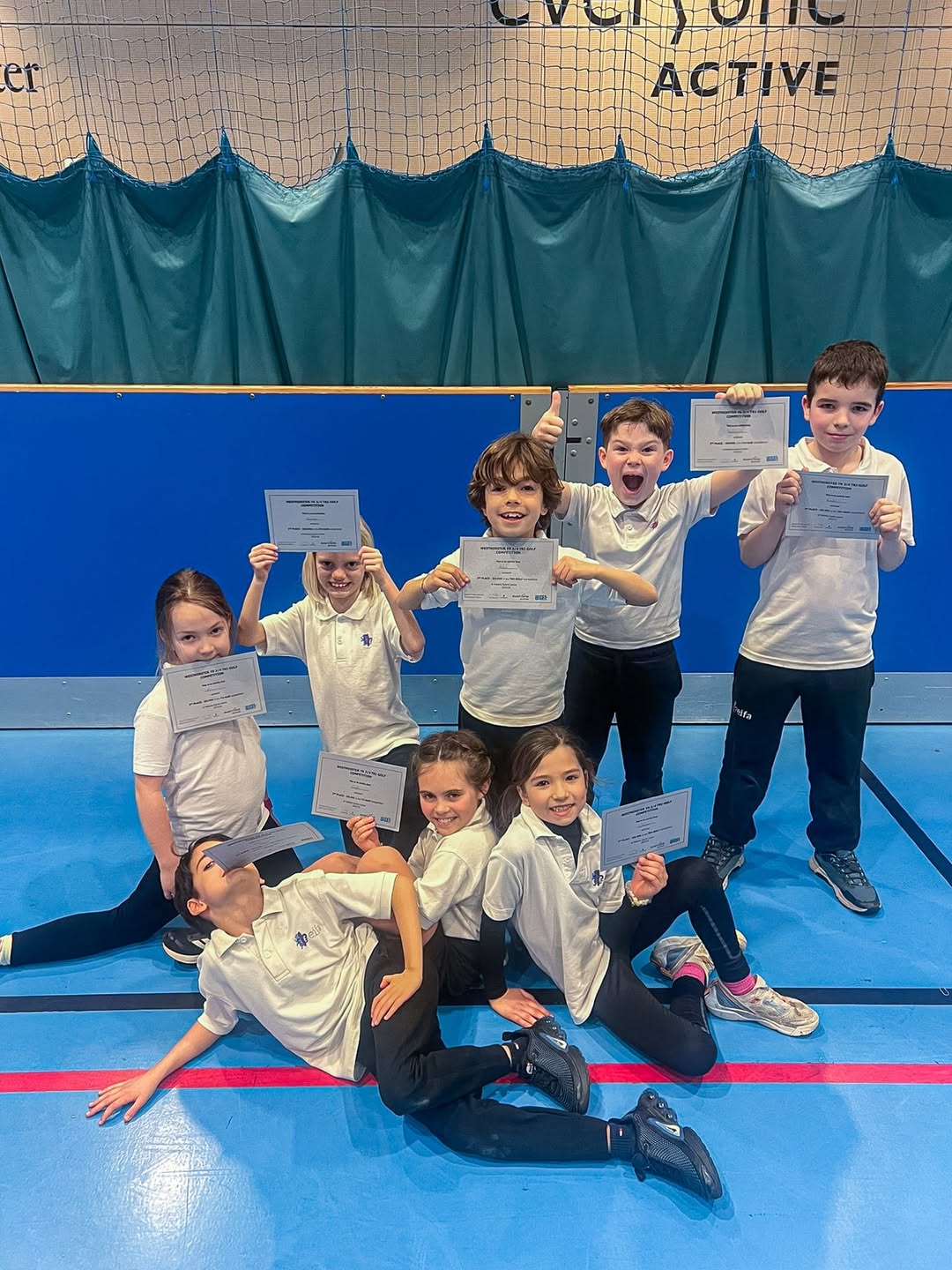EIFA Huit enfants en tenue de sport posent sur le sol d'un gymnase, tenant des certificats et souriant fiÚrement. DerriÚre eux, un filet de volley-ball et un rideau bleu mettent en scÚne leur réussite, célébrée lors de la Journée mondiale de l'alimentation.