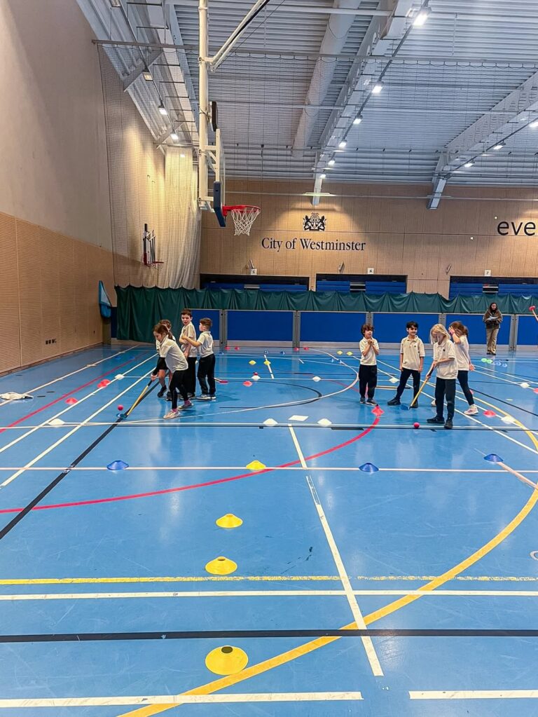 EIFA Un groupe d'enfants en tenue de sport joue avec des bùtons et des ballons sur un terrain de sport intérieur bleu, entouré de cÎnes et de lignes. Sur le mur à l'arriÚre-plan, on peut lire City of Westminster, qui célÚbre la Journée mondiale de l'alimentation en s'amusant.
