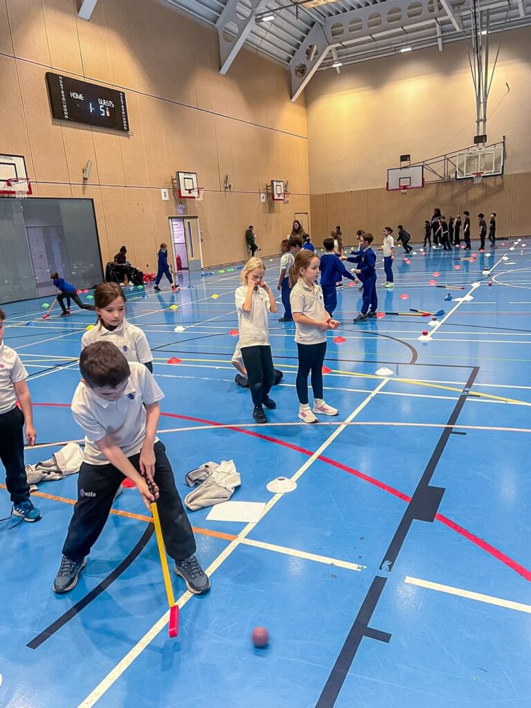 EIFA Des enfants en tenue de sport jouent avec des ballons et des battes en plastique dans un grand gymnase couvert pendant la Journée mondiale de l'alimentation. D'autres groupes sont alignés à l'arriÚre-plan et des cÎnes colorés délimitent des zones d'activité sur le sol bleu.