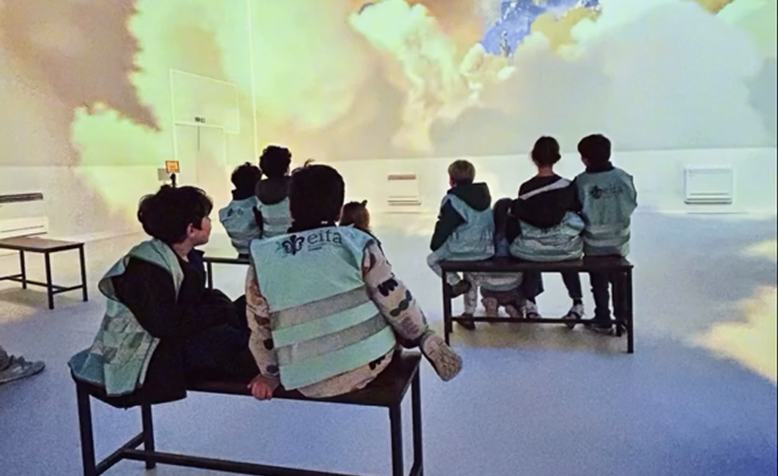 EIFA Un groupe d'enfants portant des gilets bleu clair assortis sont assis sur des bancs, face à un mur avec une grande projection immersive de nuages et de ciel dans une salle très éclairée, célébrant la Journée mondiale de l'alimentation.