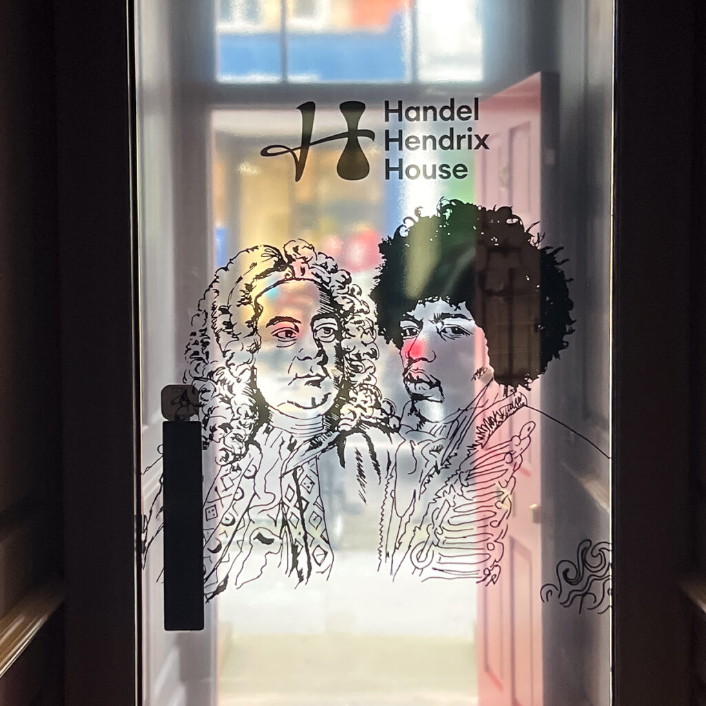 EIFA Une illustration au trait de deux personnages historiques orne une porte vitrée sous le texte « Handel Hendrix House » à Londres. Le design bilingue ajoute de la profondeur lorsque la porte souvre sur une vue animée de la rue, où les bâtiments flous contrastent avec lœuvre dart détaillée.