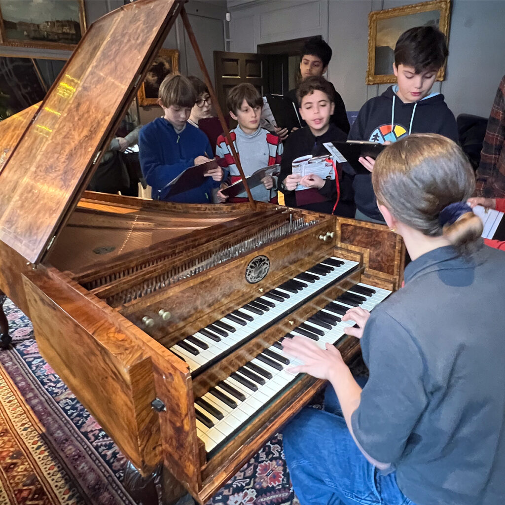 EIFA Un groupe de jeunes se rassemble autour dun professeur jouant sur un piano en bois ancien dans une école bilingue de Londres. Ils tiennent des livrets et observent attentivement dans une salle chaleureusement éclairée et ornée de tableaux encadrés sur les murs.