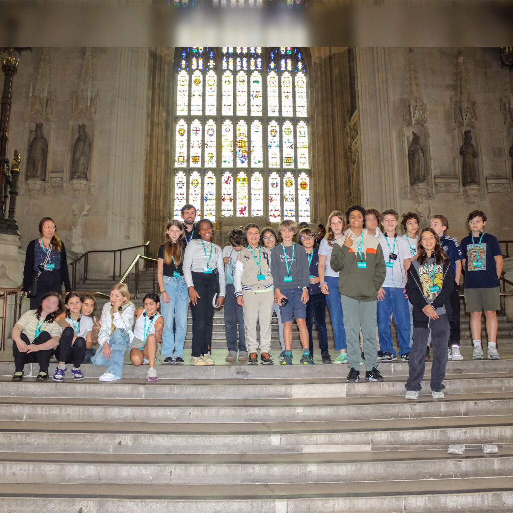 EIFA A group of teenagers, wearing lanyards and casual clothing, are gathered on steps inside a historic building in London. The large stained glass windows behind them cast colorful patterns, creating a vibrant backdrop for the students from EIFA as they engage in their school trip adventure.