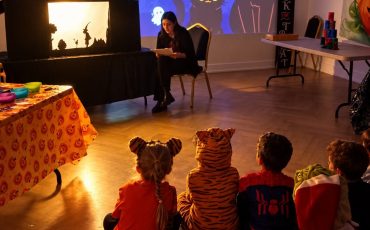 EIFA A woman performs a shadow puppet show for children in Halloween costumes at an International Open Day, with a large Happy Halloween projection on the wall behind them.