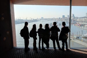 EIFA Cinq enfants portant des sacs à dos se tiennent en silhouette devant une grande fenêtre, surplombant un paysage urbain avec de l’eau, des grues et des bâtiments sous un ciel clair, réfléchissant à l’importance de la Journée mondiale de l’alimentation. EIFA Cinq enfants portant des sacs à dos se tiennent en silhouette devant une grande fenêtre, surplombant un paysage urbain avec de l'eau, des grues et des bâtiments sous un ciel clair, réfléchissant à l'importance de la Journée mondiale de l'alimentation.