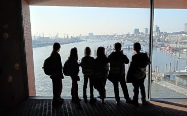 EIFA Five children wearing backpacks stand silhouetted in front of a large window overlooking a river, city buildings, and cranes on a sunny International Open Day.