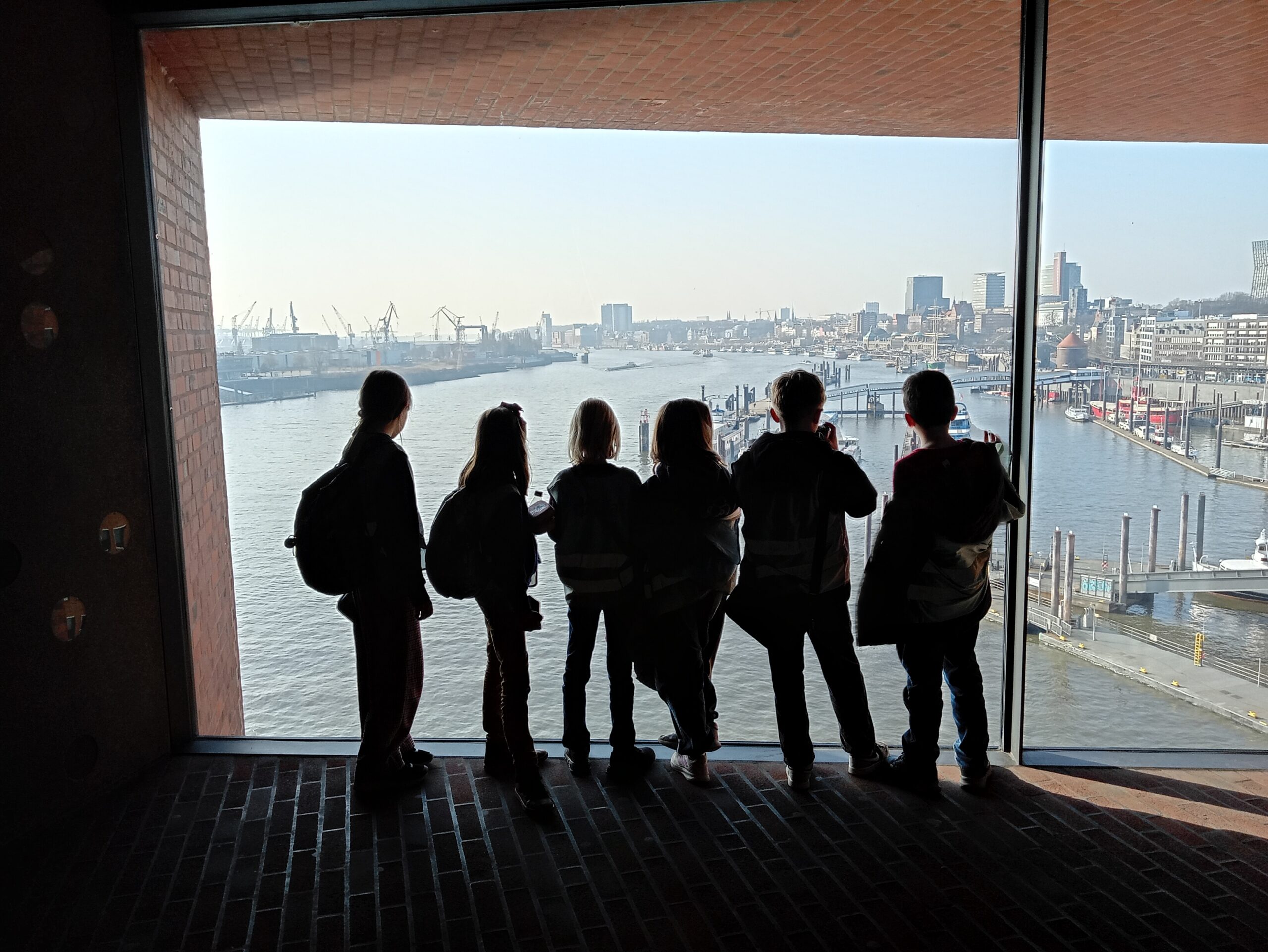 EIFA Five children wearing backpacks stand silhouetted in front of a large window overlooking a river, city buildings, and cranes on a sunny International Open Day.