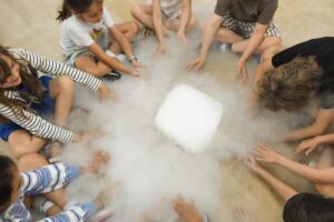 EIFA Eight children sit in a circle on a wooden floor, reaching towards a block of dry ice in the center, surrounded by misty vapor—capturing the excitement and curiosity of International Open Day. EIFA Eight children sit in a circle on a wooden floor, reaching towards a block of dry ice in the center, surrounded by misty vapor—capturing the excitement and curiosity of International Open Day.